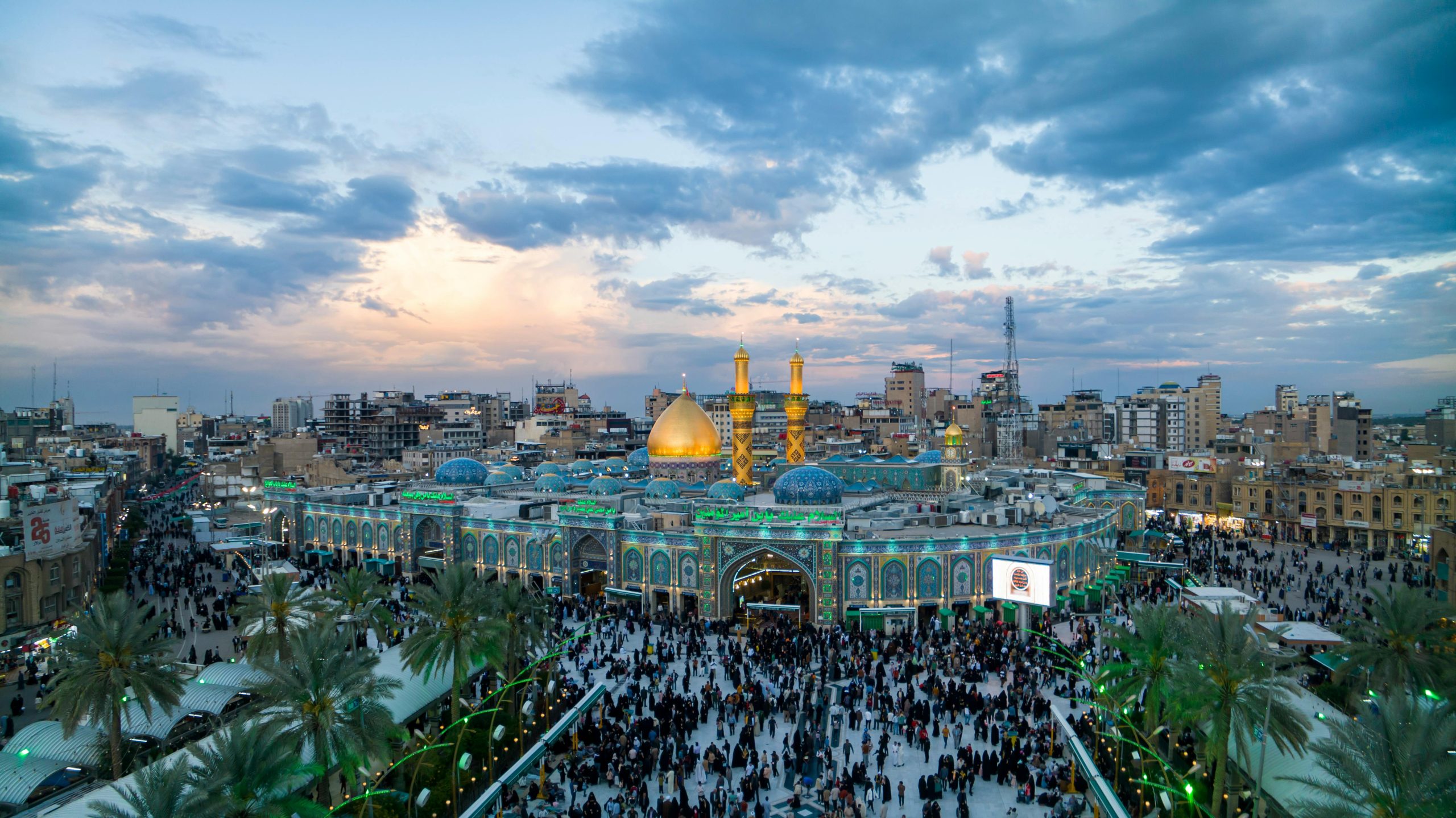 Aerial view of the Imam Husayn Shrine in Karbala, Iraq with a bustling crowd and vibrant evening sky.
